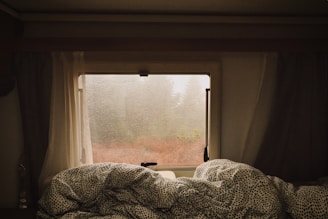 A cozy interior view of a caravan with a window showing a rainy landscape outside. Raindrops cover the glass, and the light is diffused through curtains, creating a warm ambiance. A bed with patterned blankets is visible in the foreground, adding to the sense of comfort and relaxation during a rainy day.