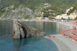 A scenic coastal area featuring a large rock formation extending into a calm sea. The shoreline is lined with numerous beach umbrellas and loungers, suggesting a popular beach destination. Vegetation-covered hills and a collection of pastel-colored buildings in the background provide a picturesque backdrop.