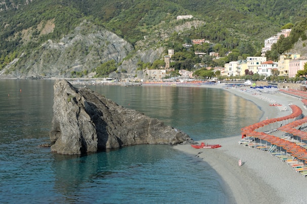 A scenic coastal area featuring a large rock formation extending into a calm sea. The shoreline is lined with numerous beach umbrellas and loungers, suggesting a popular beach destination. Vegetation-covered hills and a collection of pastel-colored buildings in the background provide a picturesque backdrop.