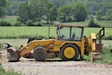A yellow tractor with a front loader attachment is parked on a gravel path with a lush, green field and trees in the background. The tractor's cabin is visible and some farming equipment can be seen nearby.