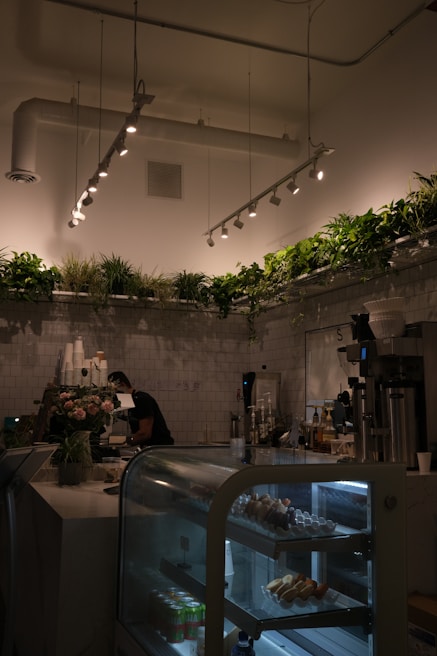 A cozy cafe interior featuring a display case with pastries and drinks, a barista working behind the counter, and abundant greenery along the top perimeter. The space is softly lit with modern ceiling lights, adding to the warm atmosphere.