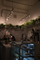 A cozy cafe interior featuring a display case with pastries and drinks, a barista working behind the counter, and abundant greenery along the top perimeter. The space is softly lit with modern ceiling lights, adding to the warm atmosphere.