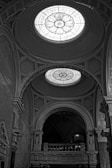 An architectural view of an ornate interior featuring large, intricately designed stained glass domes on the ceiling. The space showcases detailed molding and archways with elegant columns and artistic patterns. The monochrome setting emphasizes the textures and structural elegance of the building.