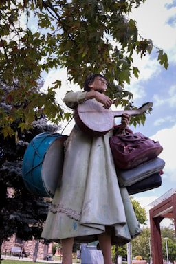 A statue of a person holding a guitar and surrounded by musical instruments, with trees and a cloudy sky in the background. The sculpture features detailed clothing and is set in an outdoor environment.