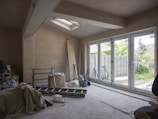 An unfinished room with exposed plaster walls and construction materials scattered around. A large ladder, tools, and bags are visible, along with covered furniture. A set of glass doors opens out to a fenced backyard with trees and grass, providing natural light into the space.