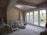 An unfinished room with exposed plaster walls and construction materials scattered around. A large ladder, tools, and bags are visible, along with covered furniture. A set of glass doors opens out to a fenced backyard with trees and grass, providing natural light into the space.