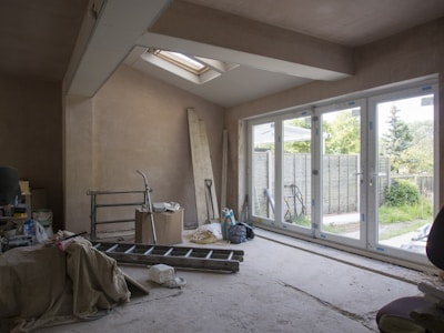 An unfinished room with exposed plaster walls and construction materials scattered around. A large ladder, tools, and bags are visible, along with covered furniture. A set of glass doors opens out to a fenced backyard with trees and grass, providing natural light into the space.