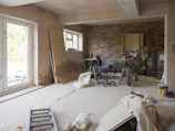 A room under renovation with exposed brick walls and bare plaster surfaces. A ladder lies on the floor, and various construction materials like wooden boards, insulation, and bags of plaster are scattered around. A chair and some tools are placed near the wall, and there's a large window allowing natural light into the space.
