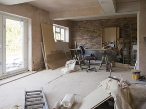 A room under renovation with exposed brick walls and bare plaster surfaces. A ladder lies on the floor, and various construction materials like wooden boards, insulation, and bags of plaster are scattered around. A chair and some tools are placed near the wall, and there's a large window allowing natural light into the space.