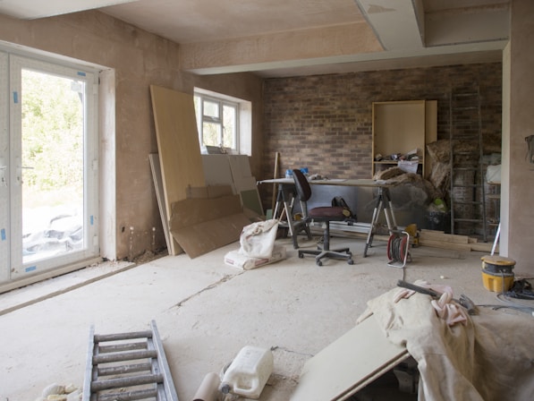 A room under renovation with exposed brick walls and bare plaster surfaces. A ladder lies on the floor, and various construction materials like wooden boards, insulation, and bags of plaster are scattered around. A chair and some tools are placed near the wall, and there's a large window allowing natural light into the space.