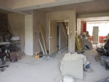A room under renovation with various construction tools and materials scattered around. There are ladders, paint cans, boards, and loose wires on the floor. Furniture is covered with protective sheets. The walls are partially unfinished, and there is a suspended ceiling. A light source from a window at the back illuminates the room.