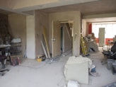 A room under renovation with various construction tools and materials scattered around. There are ladders, paint cans, boards, and loose wires on the floor. Furniture is covered with protective sheets. The walls are partially unfinished, and there is a suspended ceiling. A light source from a window at the back illuminates the room.