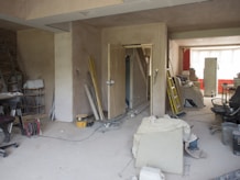 A room under renovation with various construction tools and materials scattered around. There are ladders, paint cans, boards, and loose wires on the floor. Furniture is covered with protective sheets. The walls are partially unfinished, and there is a suspended ceiling. A light source from a window at the back illuminates the room.