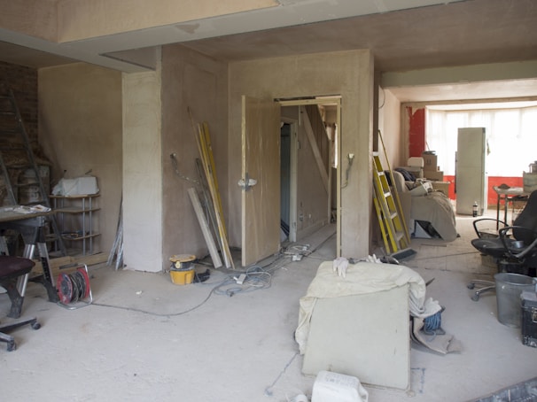 A room under renovation with various construction tools and materials scattered around. There are ladders, paint cans, boards, and loose wires on the floor. Furniture is covered with protective sheets. The walls are partially unfinished, and there is a suspended ceiling. A light source from a window at the back illuminates the room.