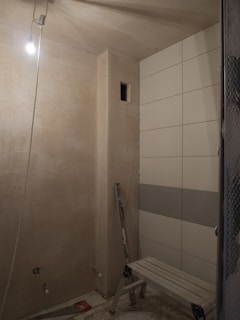 An unfinished room with bare walls and tiled section on the right. A single hanging light bulb illuminates the space, revealing construction tools such as a stepladder and a saw on the floor. The room appears to be a work in progress, possibly for a renovation or construction project.