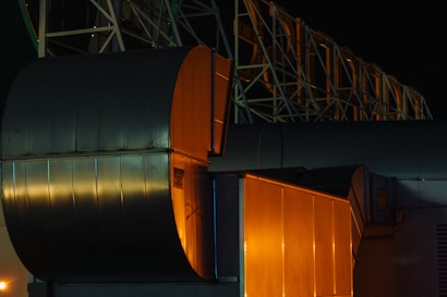 Large industrial structures with dark metallic surfaces and orange lighting. Geometric shapes and angular forms are prominent, set against a dark background. The image conveys a sense of technology or architecture.
