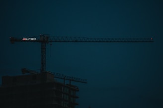 A dark construction site features a large, illuminated crane with the silhouette of an unfinished building beneath. The sky is a deep blue, indicating either early morning or evening lighting.