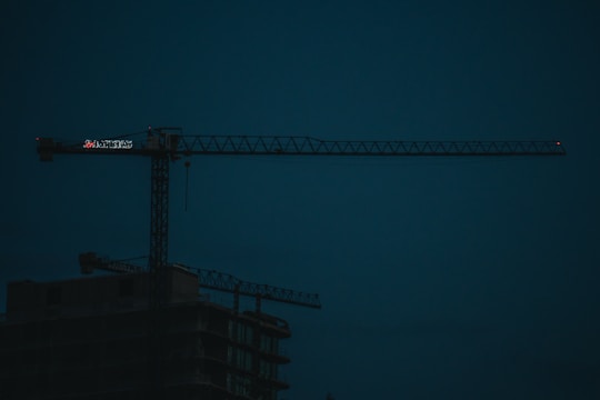 A dark construction site features a large, illuminated crane with the silhouette of an unfinished building beneath. The sky is a deep blue, indicating either early morning or evening lighting.