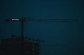 A dark construction site features a large, illuminated crane with the silhouette of an unfinished building beneath. The sky is a deep blue, indicating either early morning or evening lighting.