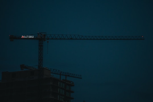 A dark construction site features a large, illuminated crane with the silhouette of an unfinished building beneath. The sky is a deep blue, indicating either early morning or evening lighting.