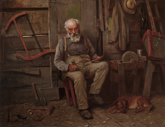 An elderly man with a white beard sits in a rustic cabin, holding a small object in his hand. He is dressed in casual work clothes and rests on a wooden chair. Around him are various tools, a hat hanging on the wall, and a sleeping dog lying on the wooden floor. The setting gives a sense of an old workshop or a shed.