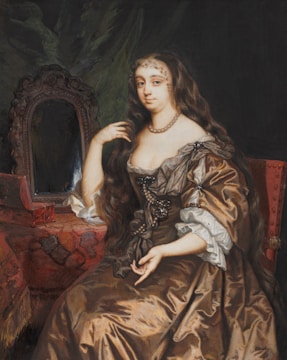 A woman dressed in historical attire is seated, surrounded by luxurious fabrics. Her dress is an elaborate brown gown with ornate details, and she wears a pearl necklace. Her hair is long and cascading over her shoulders. Beside her is a decorative mirror with a richly carved frame, reflecting her image, and a jewelry box sits on the draped table.