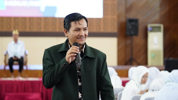A person in a green jacket holds a microphone, standing in front of an audience. The background features a seated individual and people dressed in white attire, suggesting a formal or ceremonial setting.