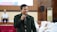 A person in a green jacket holds a microphone, standing in front of an audience. The background features a seated individual and people dressed in white attire, suggesting a formal or ceremonial setting.