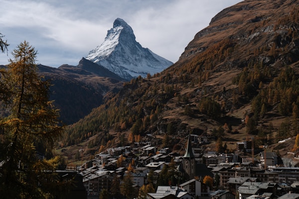 A dramatic mountain landscape features a prominent, snow-capped peak rising sharply into a partly cloudy sky. Below, a village with traditional alpine architecture nestles in a valley surrounded by rugged, forested hills that display autumnal colors. The scene conveys a sense of grandeur and tranquility.
