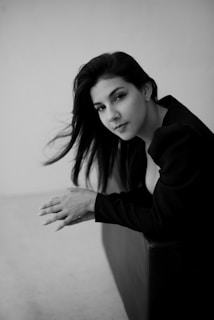A monochrome portrait of a woman with long dark hair, wearing a black outfit, leaning on a surface with her hands clasped. Her hair is gently blowing, and she is gazing towards the camera with a neutral expression.