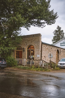 A rustic stone building with arched windows is situated amidst large trees. The front is adorned with a wooden railing and signage, and the road in front is wet, suggesting recent rain. Two cars are parked nearby, enhancing the quaint, serene atmosphere.
