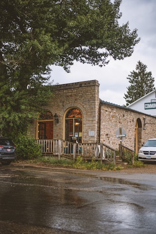 A rustic stone building with arched windows is situated amidst large trees. The front is adorned with a wooden railing and signage, and the road in front is wet, suggesting recent rain. Two cars are parked nearby, enhancing the quaint, serene atmosphere.