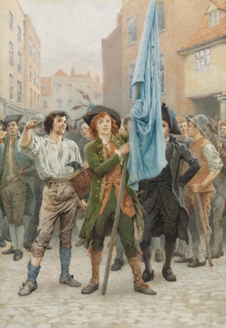 A group of people, primarily men, gather in a cobblestone street in front of historic buildings. The central figure holds a large blue flag with an assertive expression. The clothing suggests a historical period, featuring breeches, waistcoats, and tricorn hats. The surrounding individuals appear animated and engaged in what seems like a demonstration or rally.