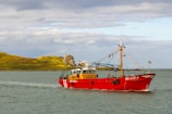 A bright red fishing boat is sailing on a calm body of water with a small, rocky green island in the background. The sky is partly cloudy, and the sunlight is casting a warm glow on the scene. The boat has visible equipment and rigging for fishing.