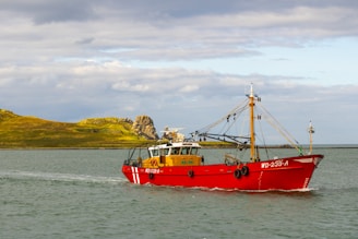 A bright red fishing boat is sailing on a calm body of water with a small, rocky green island in the background. The sky is partly cloudy, and the sunlight is casting a warm glow on the scene. The boat has visible equipment and rigging for fishing.