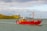 A bright red fishing boat is sailing on a calm body of water with a small, rocky green island in the background. The sky is partly cloudy, and the sunlight is casting a warm glow on the scene. The boat has visible equipment and rigging for fishing.