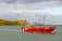 A bright red fishing boat is sailing on a calm body of water with a small, rocky green island in the background. The sky is partly cloudy, and the sunlight is casting a warm glow on the scene. The boat has visible equipment and rigging for fishing.