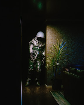 An astronaut in a space suit stands in a dimly lit indoor corner. The background features patterned wallpaper and a potted plant beside the figure. A couch partially enters the frame at the bottom right, casting subtle shadows on polished wooden flooring.