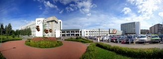 A large, modern building complex with multiple levels and a mostly white facade is situated in an urban setting. The foreground features a well-maintained brick pathway edged with neatly trimmed green hedges and a circular flower bed. Several tall lamp posts with hanging flower baskets add a decorative element. In the background, numerous parked cars fill a parking lot adjacent to the buildings. Tall trees line part of the left side, providing a natural touch. The sky appears bright and clear, with scattered clouds.