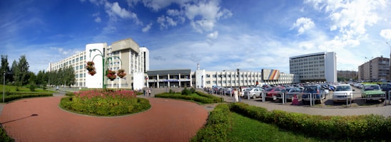 A large, modern building complex with multiple levels and a mostly white facade is situated in an urban setting. The foreground features a well-maintained brick pathway edged with neatly trimmed green hedges and a circular flower bed. Several tall lamp posts with hanging flower baskets add a decorative element. In the background, numerous parked cars fill a parking lot adjacent to the buildings. Tall trees line part of the left side, providing a natural touch. The sky appears bright and clear, with scattered clouds.