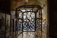 An intricately designed wrought iron gate with organic, fluid shapes is set within an arched entryway. Light filters through the openings, casting patterns on the stone walls and floor. On the left, there is a decorative glass panel showcasing floral motifs, and above hangs an ornate lantern.