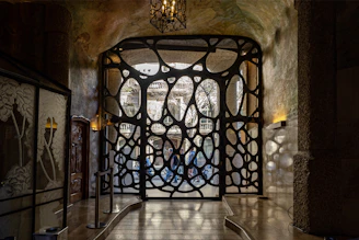 An intricately designed wrought iron gate with organic, fluid shapes is set within an arched entryway. Light filters through the openings, casting patterns on the stone walls and floor. On the left, there is a decorative glass panel showcasing floral motifs, and above hangs an ornate lantern.