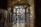 An intricately designed wrought iron gate with organic, fluid shapes is set within an arched entryway. Light filters through the openings, casting patterns on the stone walls and floor. On the left, there is a decorative glass panel showcasing floral motifs, and above hangs an ornate lantern.