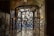 An intricately designed wrought iron gate with organic, fluid shapes is set within an arched entryway. Light filters through the openings, casting patterns on the stone walls and floor. On the left, there is a decorative glass panel showcasing floral motifs, and above hangs an ornate lantern.