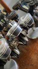Several metallic pistons are arranged on a wooden surface with reflections of light visible on their polished surfaces. Each piston is mounted on a small base, showcasing their detailed design and mechanical components.