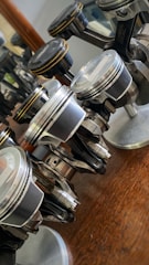 Several metallic pistons are arranged on a wooden surface with reflections of light visible on their polished surfaces. Each piston is mounted on a small base, showcasing their detailed design and mechanical components.