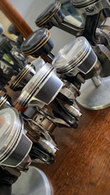 Several metallic pistons are arranged on a wooden surface with reflections of light visible on their polished surfaces. Each piston is mounted on a small base, showcasing their detailed design and mechanical components.