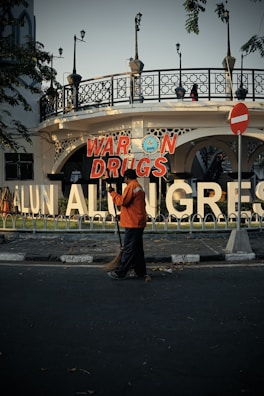 A person wearing an orange jacket is sweeping the street in front of a large signboard with the text 'WAR ON DRUGS'. The background features an ornate building with decorative iron railings, lampposts, and trees providing shade.