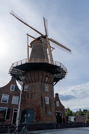 A traditional Dutch windmill with large sails standing tall against a partly cloudy sky. The windmill has a brown, brick base with multiple windows and a wooden gallery near the top. Below, there are attached buildings with dark green doors and windows. A bicycle is parked in front of the windmill, and a street lamp is visible on the left side.