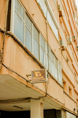 An exterior view of a tall building with large windows and multiple air conditioning units attached to the facade. There is a signboard reading 'Tabacos' with an illustration of a tobacco plant. The building's exterior is painted in a peach color, and several electrical cables are visible running along the walls.